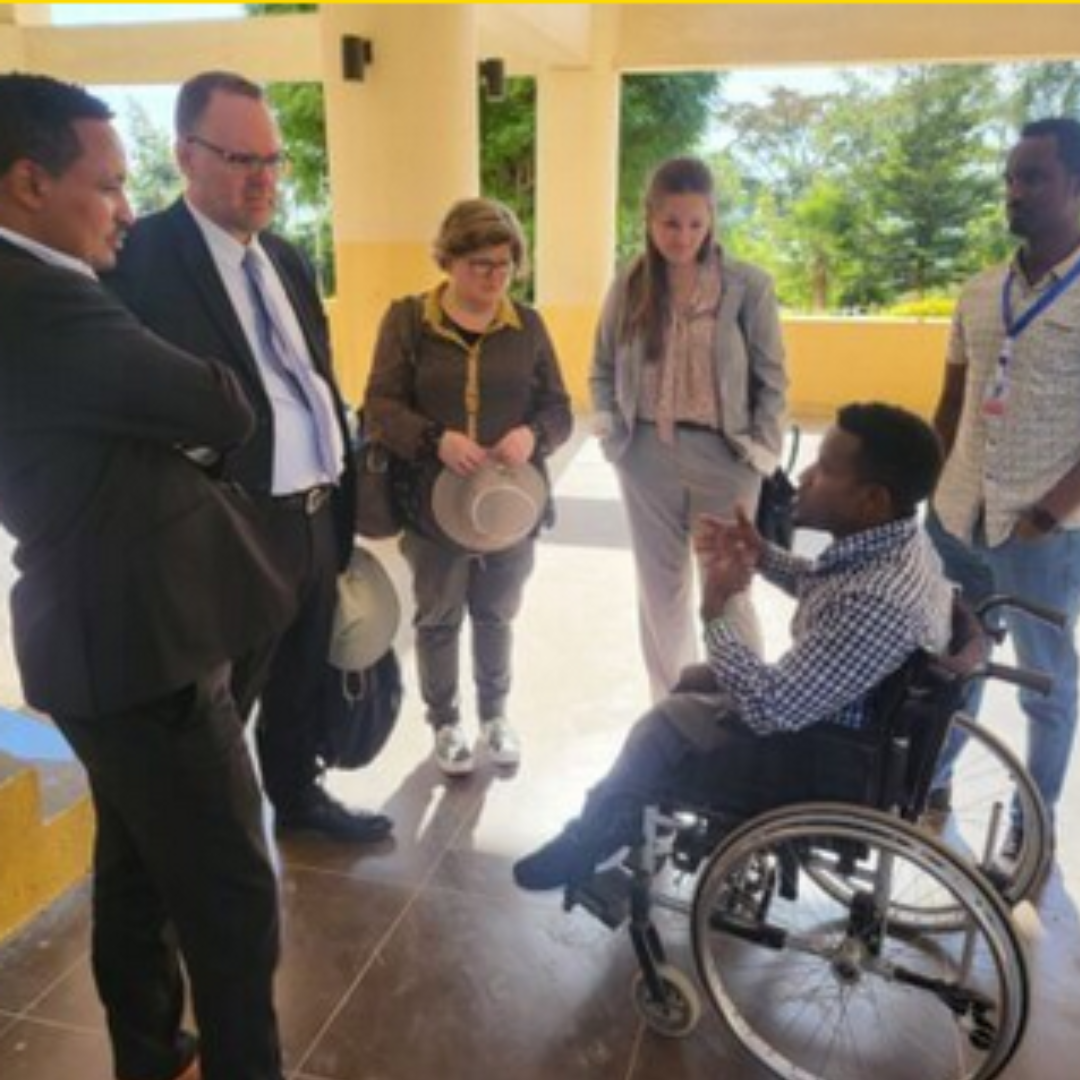 A diverse group of individuals gathered around a young Ethiopian man in a wheelchair, listening to him speak as he gestures with his hands 